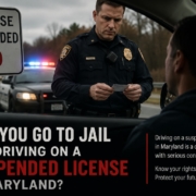 Maryland police officer conducting a traffic stop with flashing patrol lights while checking a driver’s license beside a “License Suspended MD” sign and blog headline about jail time for driving on a suspended license.