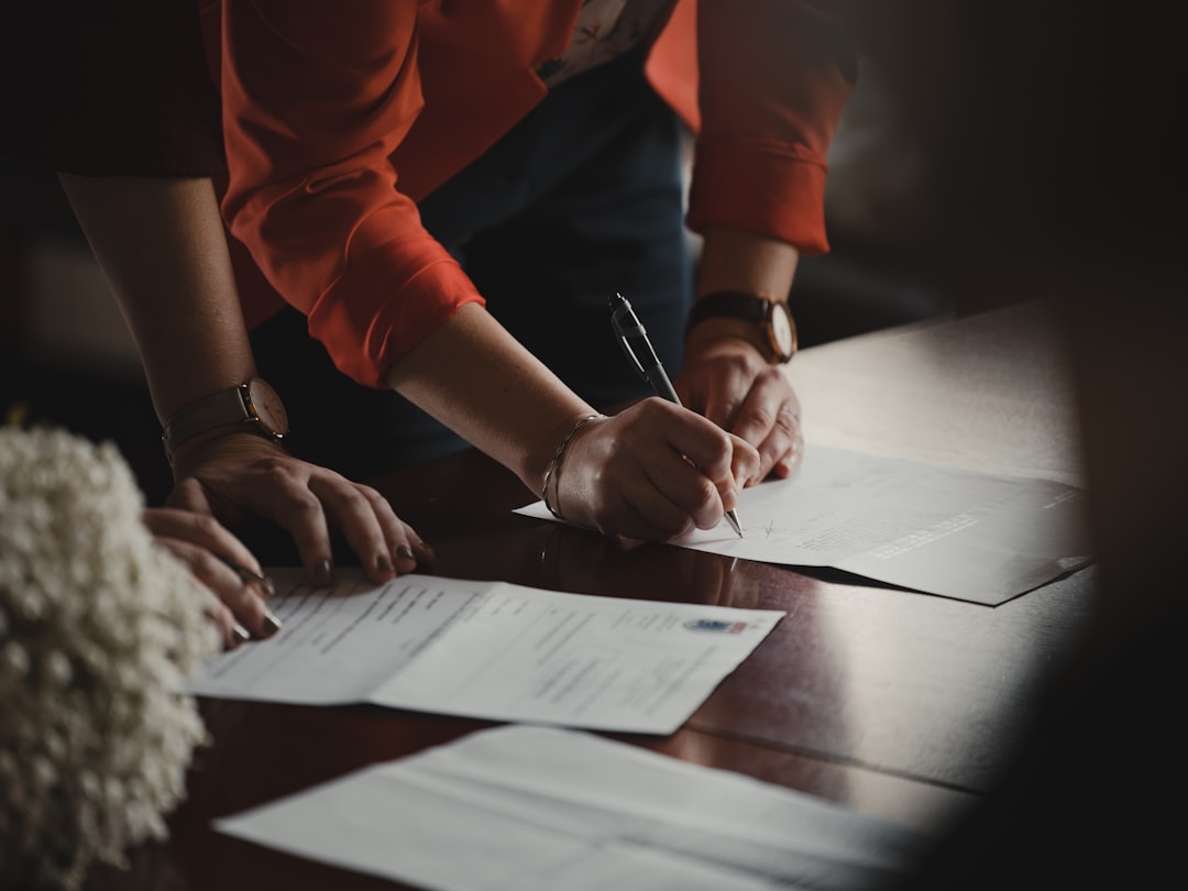 Person signing legal documents at a table while another person reviews paperwork nearby.
